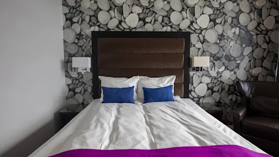Quality Hotel Ulstein rum med en bekväm säng, blå kuddar och ett lila överkast, mot en unik vägg med stenmönster.