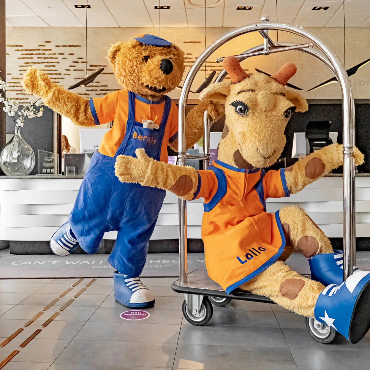 En björn och en giraff i färgglada kostymer poserar lekfullt på en bagagevagn i en hotellobby. Björnens kostym står "Bernie" och giraffens "Lollo."