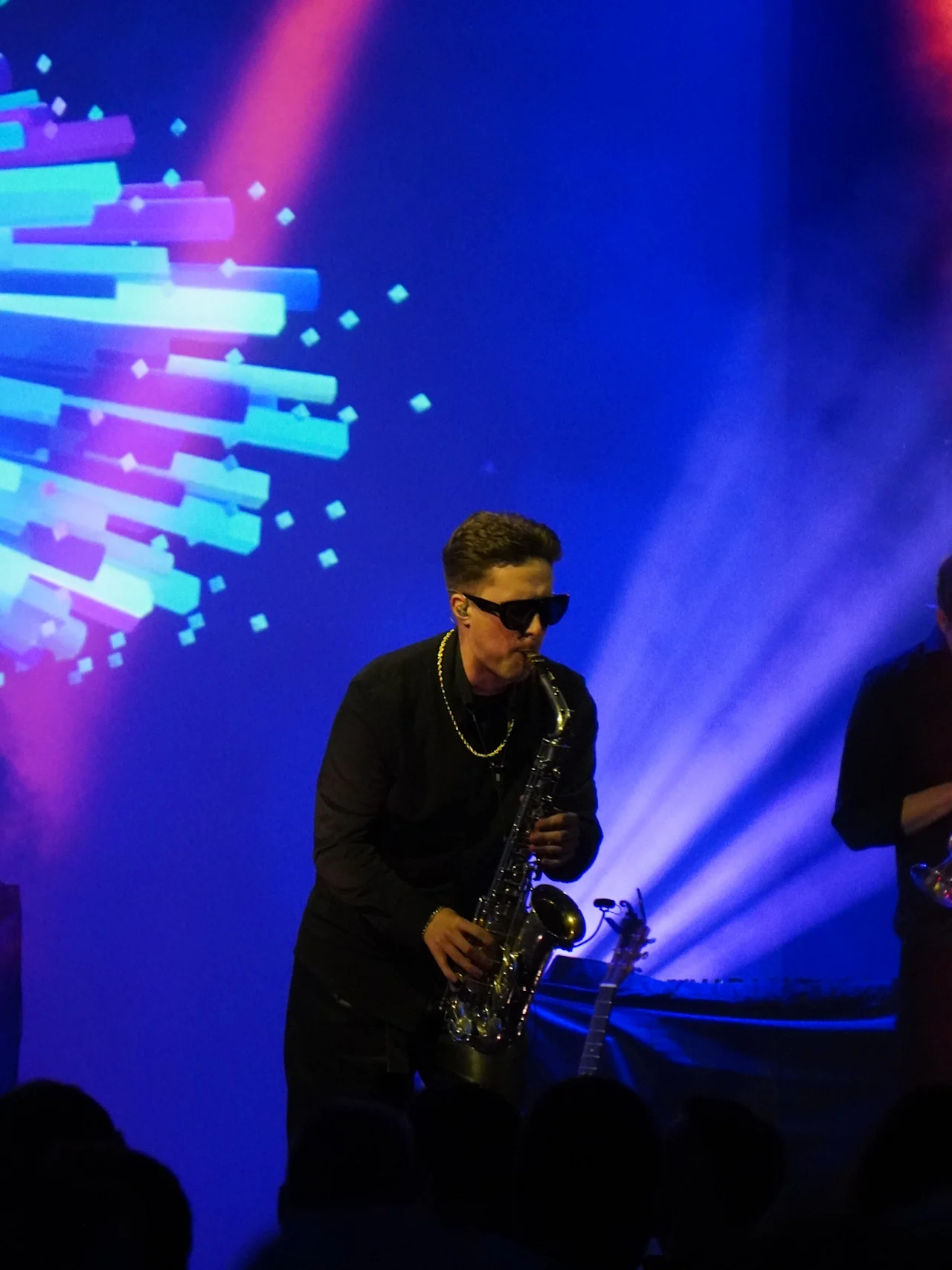 Musician playing a saxophone in front of a vibrant stage with blue and red lights, surrounded by abstract digital projections.