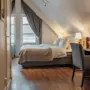 A cozy hotel room with a comfortable bed, a desk, and a chair, featuring a sloped ceiling and a window with curtains.