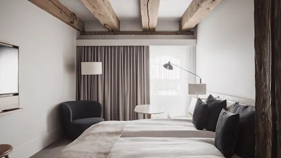 A cozy hotel room features a twin bed with dark pillows, a black chair, and a wooden table, set under rustic wooden ceiling beams.