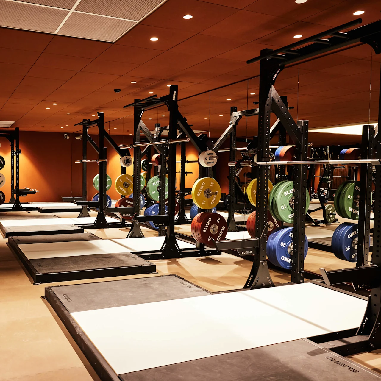 Et veludstyret fitnessrum med flere squat racks og vægtskiver inde i et rum med orange og trætoner.
