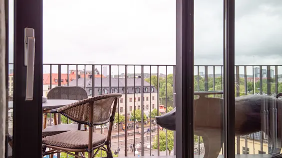 En balkong med utemöbler med utsikt över en stad med byggnader, träd och en flod under en molnig himmel. En grisstaty är också synlig.