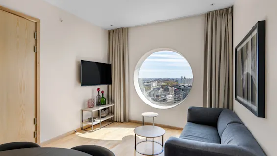 Moderne stue med et rundt vindue, der tilbyder panoramaudsigt over byen, med en komfortabel sofa og et TV.