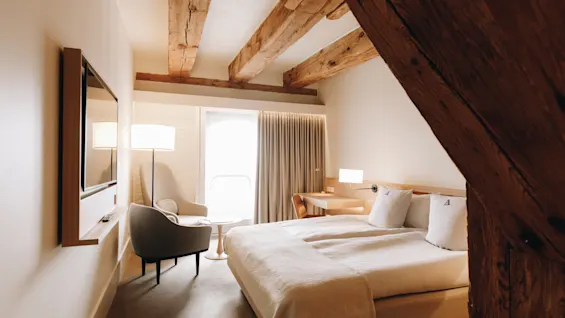 Ett mysigt hotellrum med en stor säng, synliga takbjälkar i trä, en väggmonterad TV och ett ljust fönster.