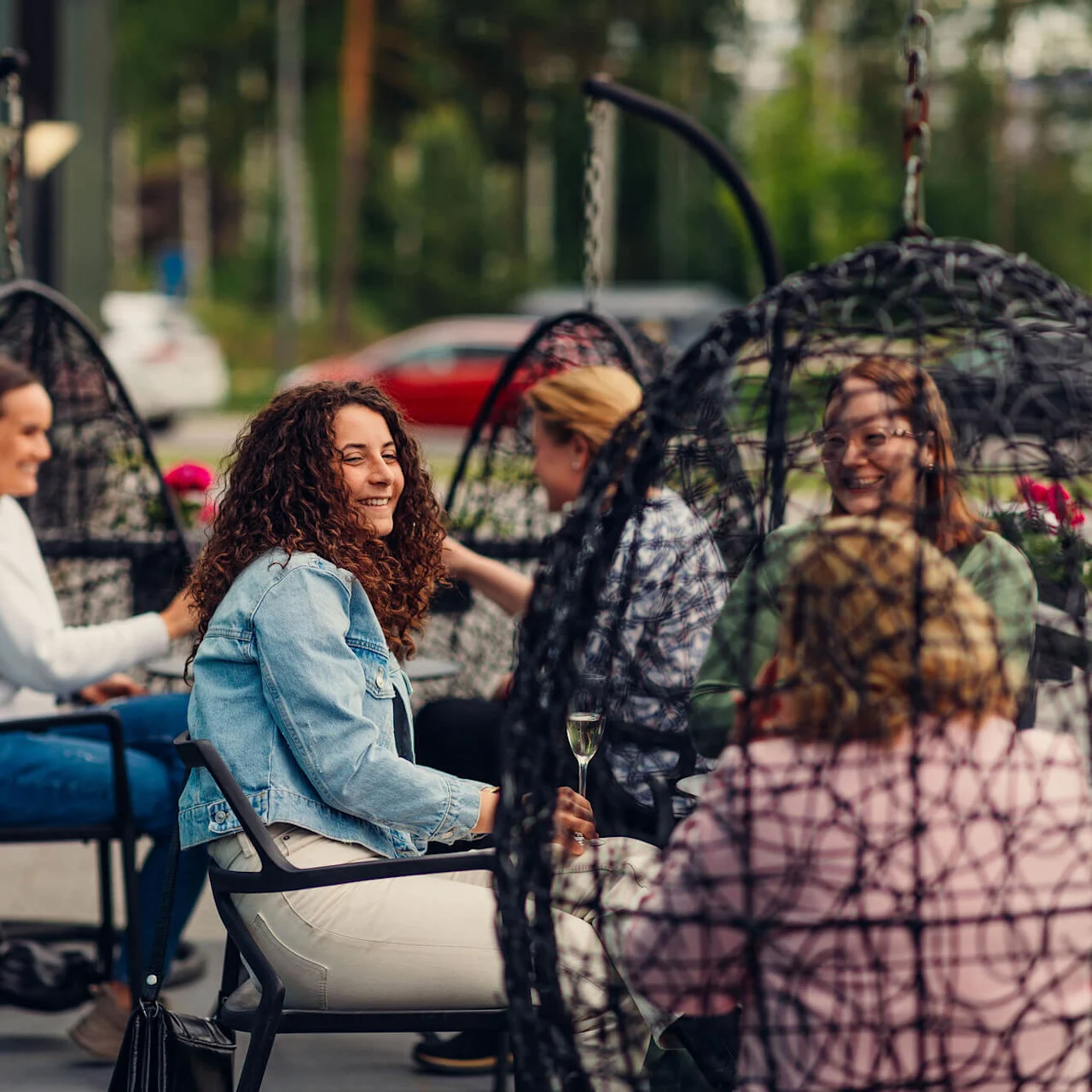 Group of women sitting and chatting in hanging chairs outdoors. They are smiling, surrounded by greenery and parked cars, suggesting a casual social gathering in a garden-like setting.
