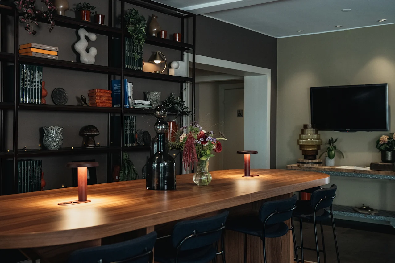 Ett varmt och inbjudande gemensamt utrymme med ett långt träbord, barstolar och glödande lampor, mot en bokhylla.