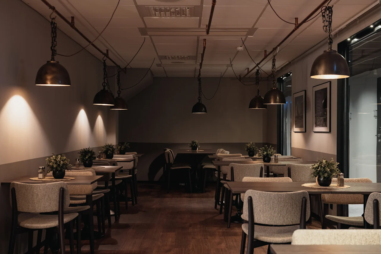 A cozy restaurant dining area with warm lighting from hanging pendant lamps, featuring multiple tables and chairs set for guests.