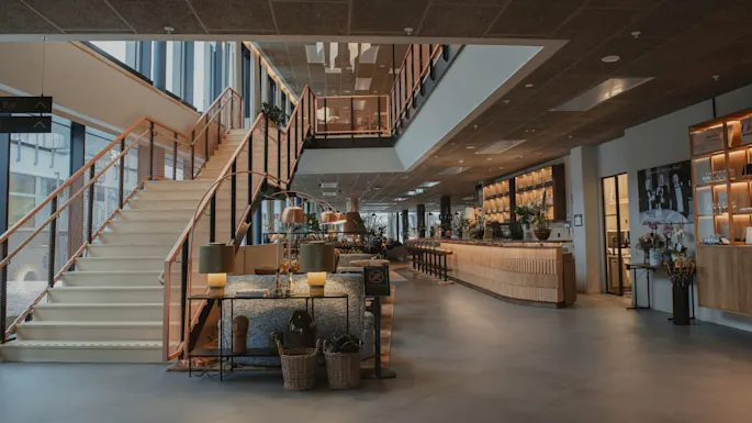 Stor trapp går opp i en moderne lobby, flankert av en elegant bar, trehyller og sitteområde. Myk belysning og planter forsterker den innbydende atmosfæren.