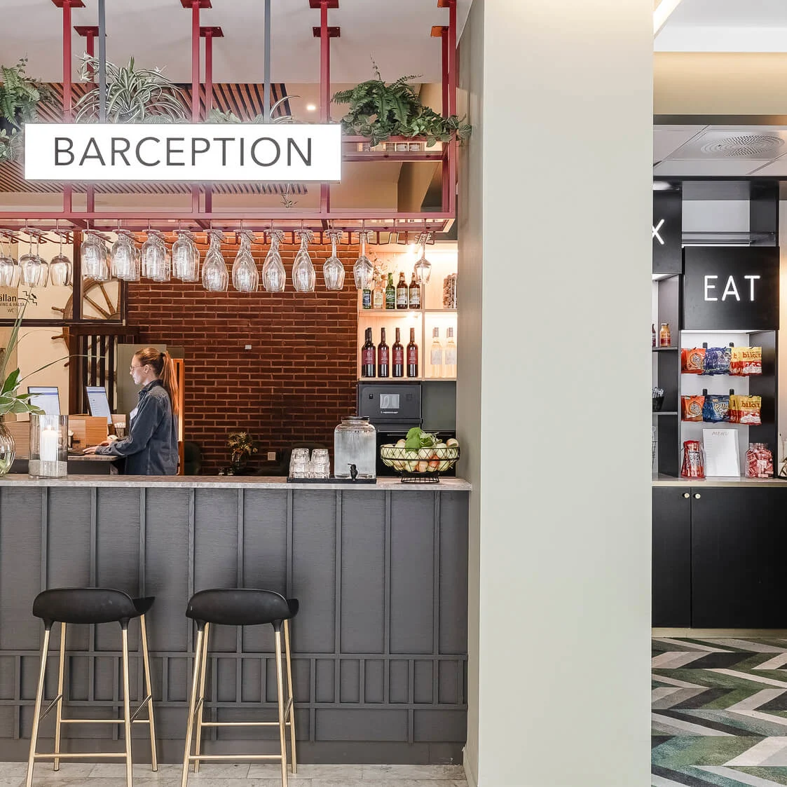 En hybrid bar och reception märkt "BARCEPTION". En person arbetar bakom den, i en välbelyst, modern lobby med växter och geometriska golvmonster.