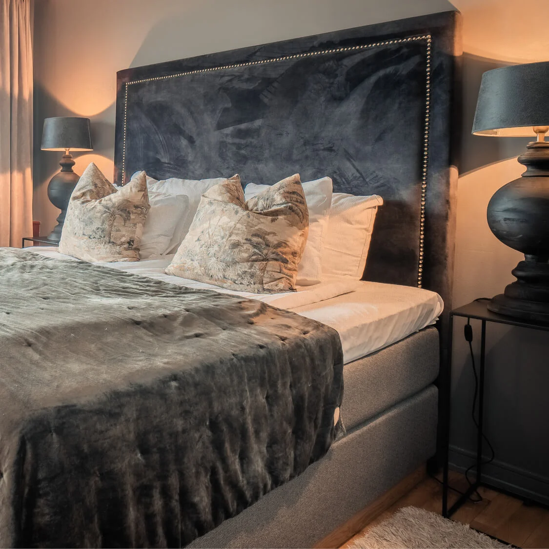 A bed with gray bedding is neatly arranged, topped with decorative pillows, situated in a warmly lit bedroom with wooden floors and a large window showing an urban view.