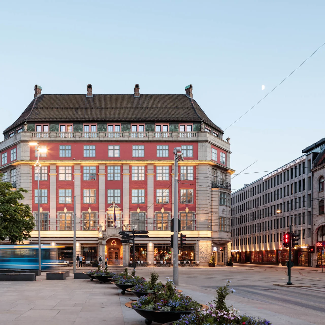 A large, ornate building stands in a city square at dusk, with a tram passing by. Text reads: "CLARION HOTEL THE HUB OSLO CENTRAL STATION." People are sitting nearby.