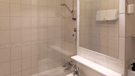 Badkar med duschmunstycke fastsatt på kaklad vägg, inneslutet av glasvägg. Ovanför badkaret, en vikt handduk på en räls. I närheten, ett handfat med kran. Neutrala färger på badrumskaklet runtom.