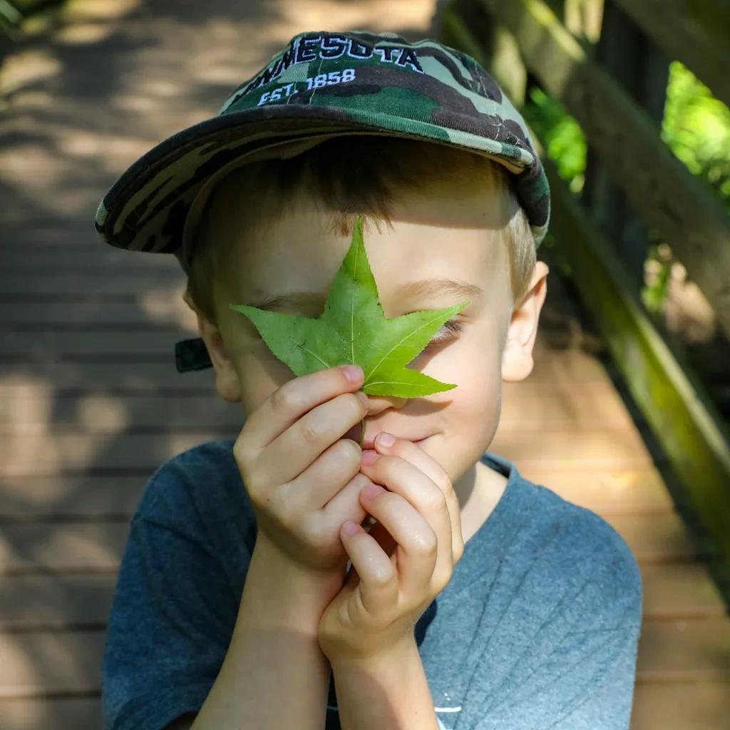 Ett barn i naturen, med en keps, håller lekfullt ett grönt löv över ansiktet på en träbro.