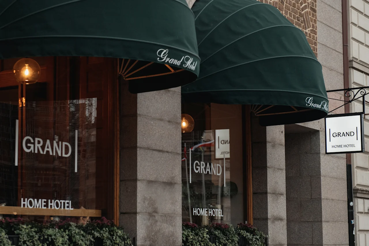 Exteriör av ett hotell med gröna markiser och skyltar som lyder Grand och Home Hotel, vilket antyder en välkomnande entré.