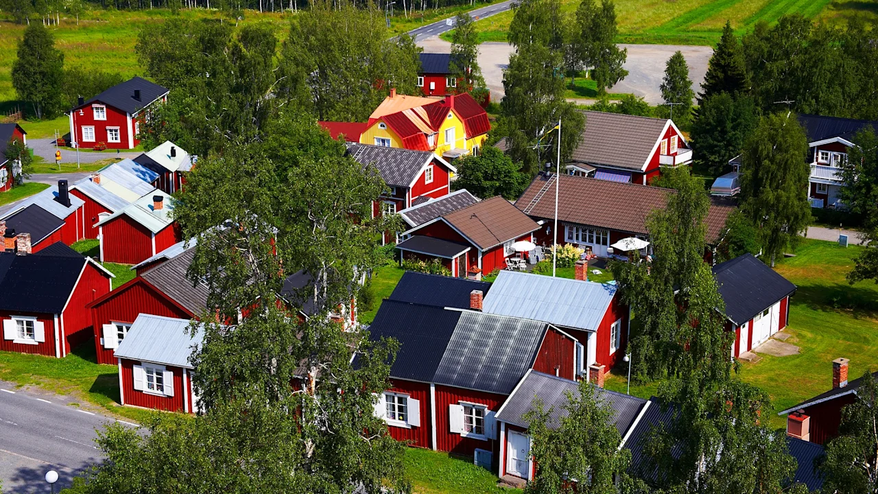 Red wooden houses with pitched roofs stand among tall green trees in a rural village on a sunny day. The scene is surrounded by fields and roads.
