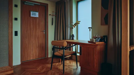 Et koselig hotellrominteriør med et skrivebord og stol ved et vindu, samt en dør med rominformasjonsskilt.