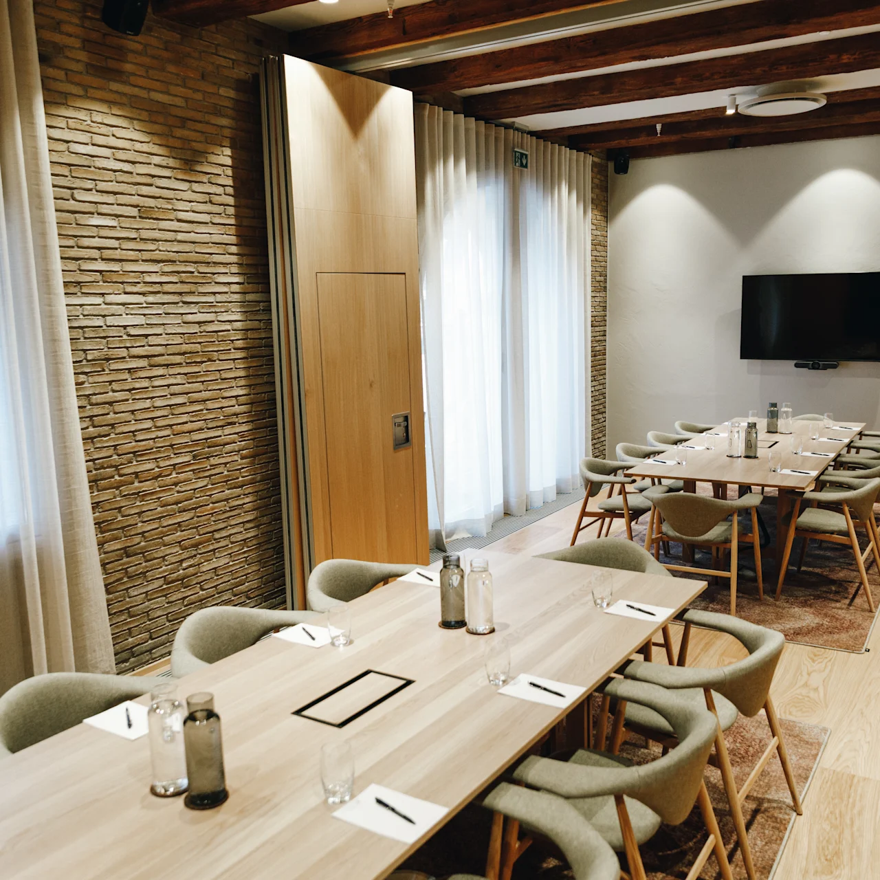Conference tables arranged with notebooks and water bottles; chairs surround the tables. A wall-mounted TV is visible. The room features brick and wood accents with soft lighting.  