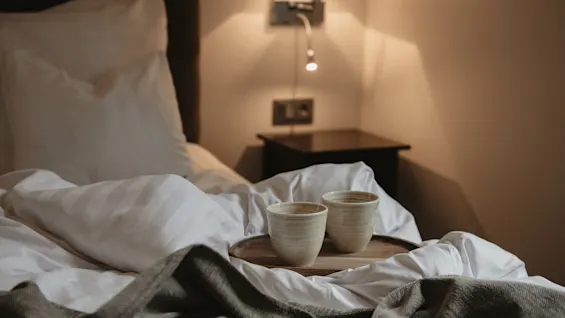 To keramiske kopper på en træbakke i en seng med hvide lagner og et gråt tæppe, hvilket antyder en hyggelig morgen på Home Hotel Norre Park i Halmstad.
