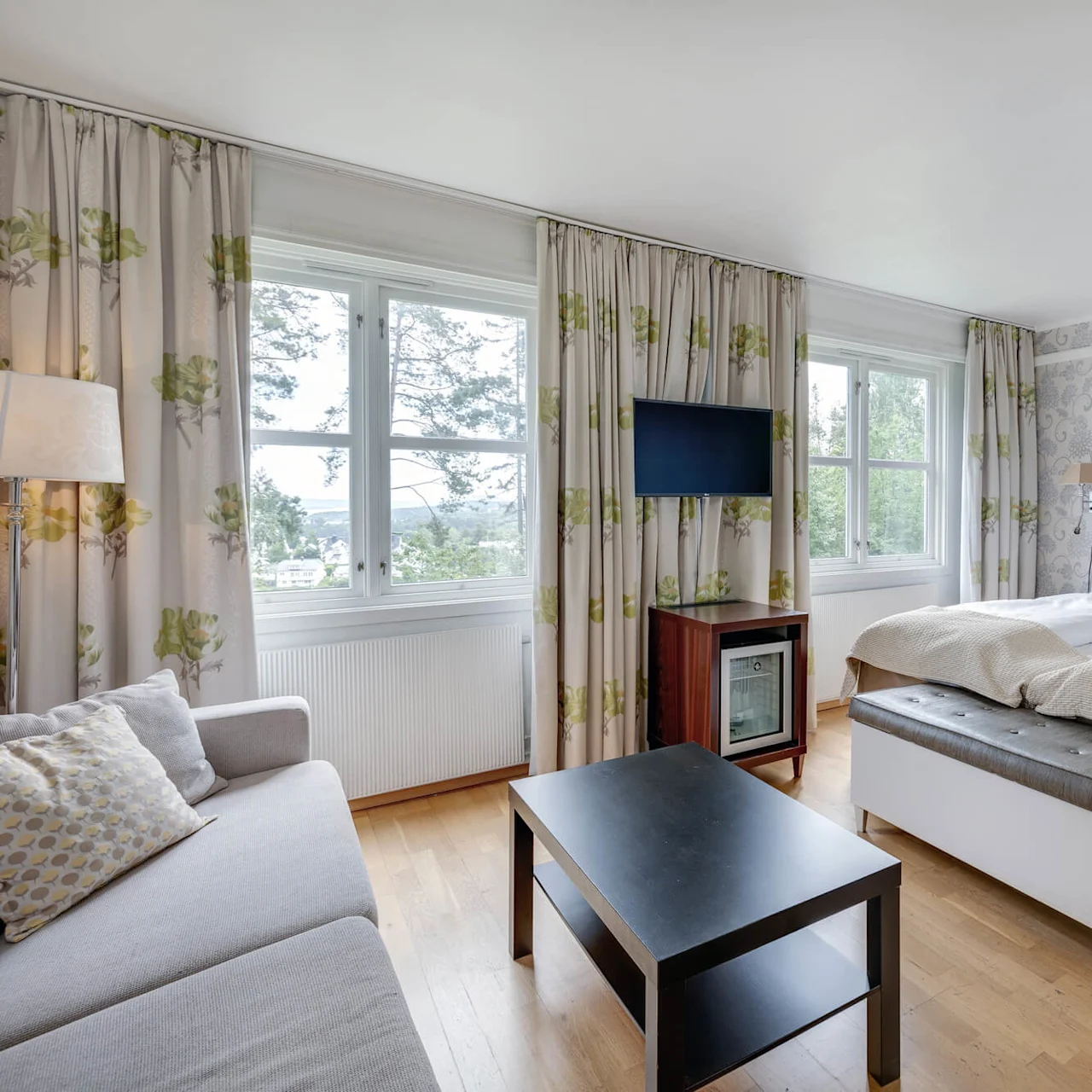 Et moderne hotellrom har to senger med dekorativt sengestøtte. Plassert nær store vinduer, inkluderer det en grå sofa, svart salongbord, TV og mønstrete gardiner.
