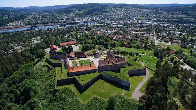 An old fortress sits amidst green hills, surrounded by a town with trees and a river in the background.