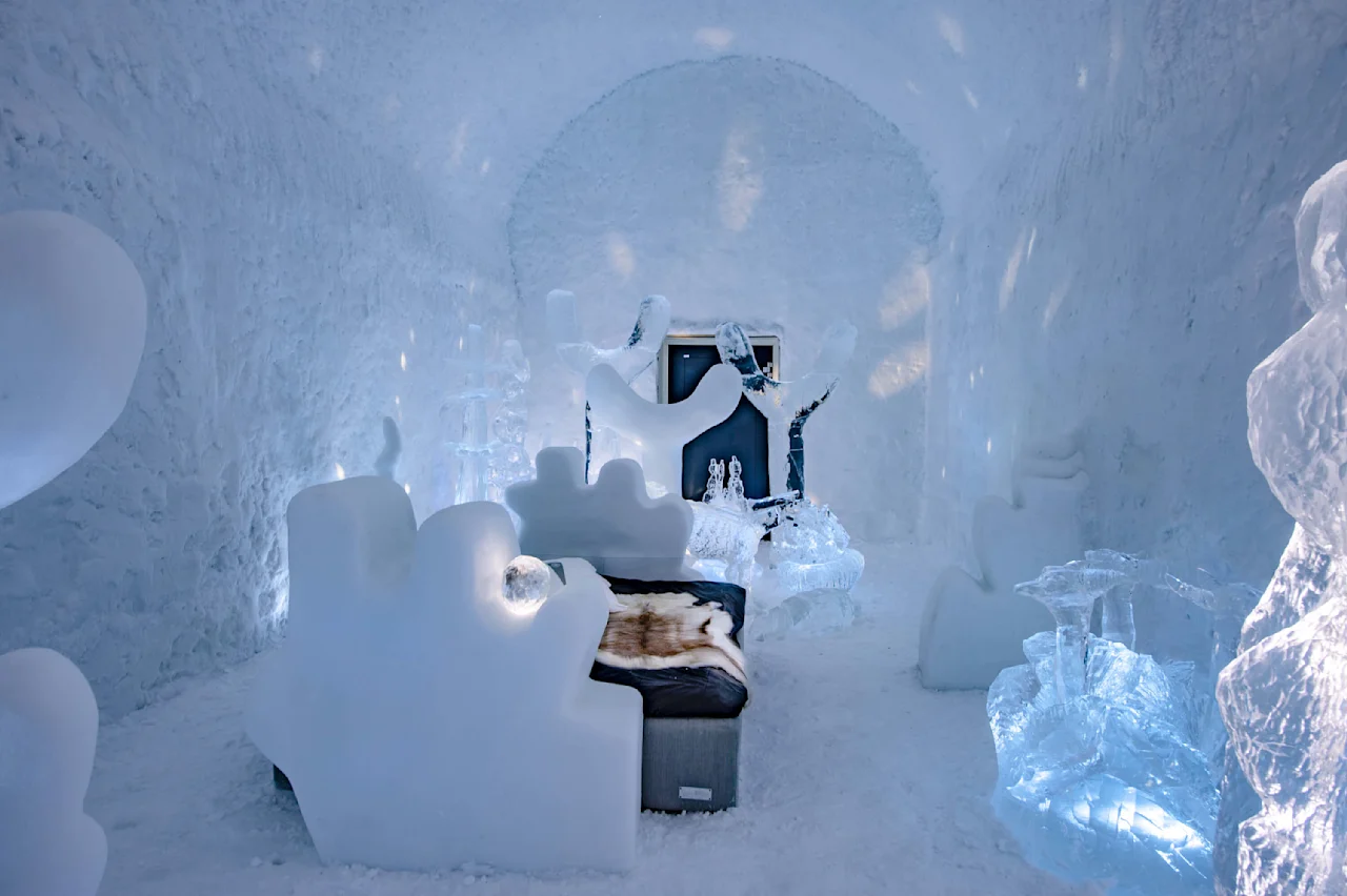 Et rum med isskulpturer har detaljeret møbler og dekorationer af is, inklusive en seng med et pelsplaid. Miljøet er oplyst med blåt lys, hvilket skaber en rolig atmosfære.