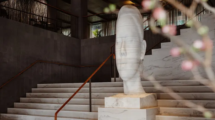 Sculpture of a serene face on marble steps, surrounded by a modern interior with dark walls and railings. Blurred pink blossoms in the foreground.