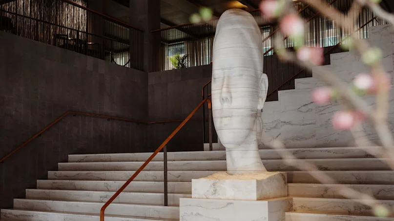 Sculpture of a serene face on marble steps, surrounded by a modern interior with dark walls and railings. Blurred pink blossoms in the foreground.