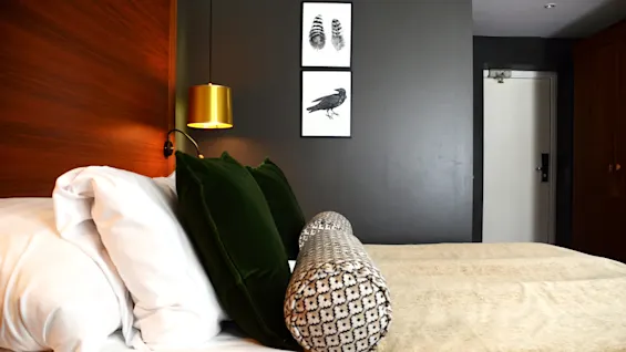 A well-made bed with plush pillows, a cylindrical cushion, and a golden lamp hanging overhead, against a backdrop of wood paneling and framed artwork, suggesting a cozy, modern bedroom setting.