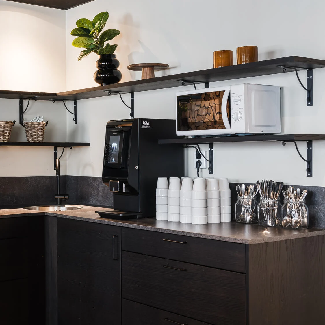 Kaffestationen har en svart kaffemaskin på en bänk med staplade koppar. Hyllor ovanför innehåller växter, glas, korgar och en mikrovågsugn, i en prydlig, modern miljö.