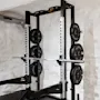 Et vægtløftestativ holder en vægtstang med forskellige vægtskiver mærket "TECHNOGYM" mod en hvid murstensvæg i et træningscenter.