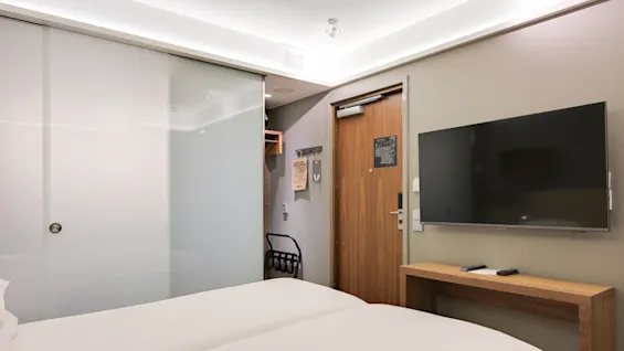 Engelska: A modern hotel room with a bed facing a mounted TV, a wooden door, and a frosted glass partition. Control remotes rest on a wooden bench.