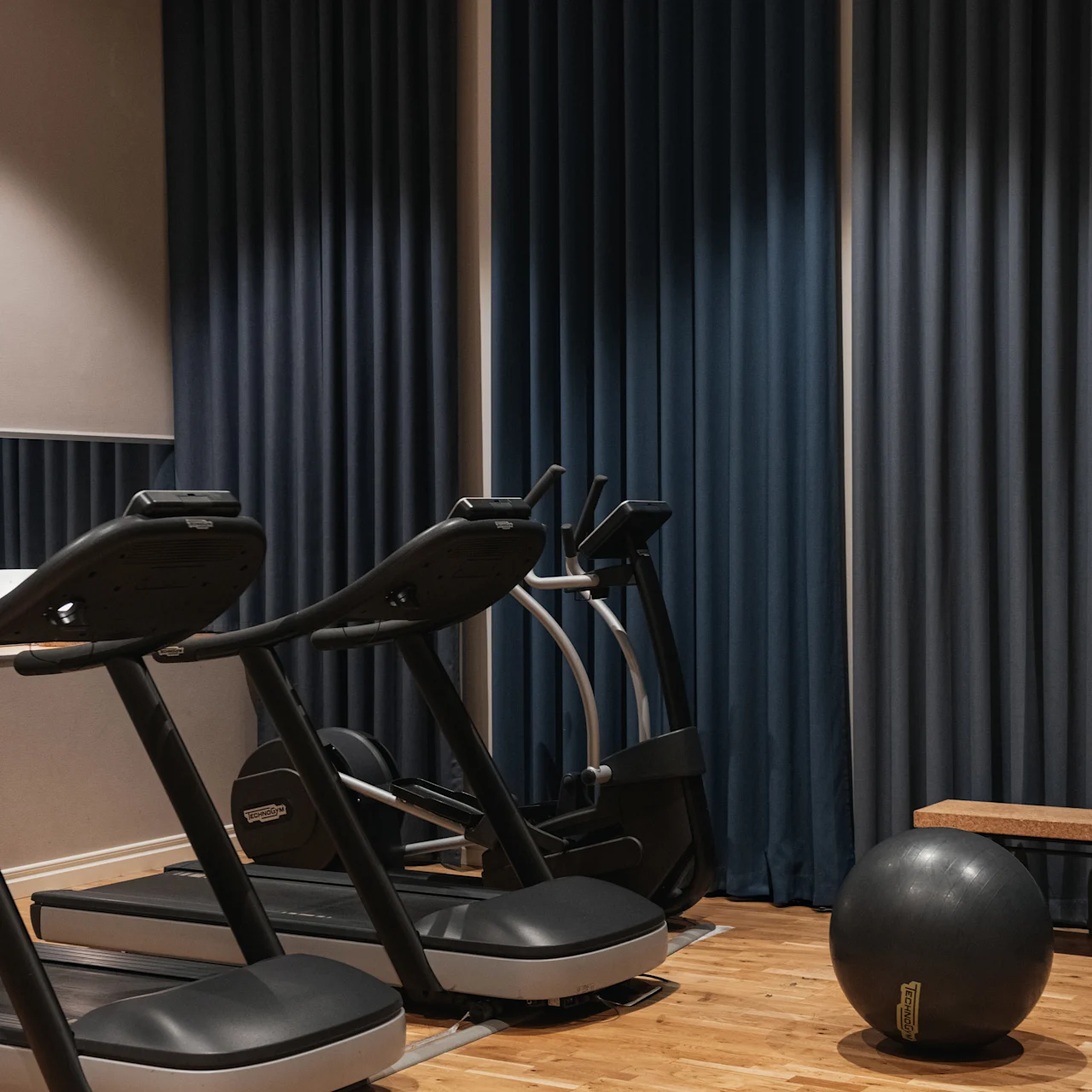 Löpband står uppradade i ett svagt upplyst gym med blå gardiner, bredvid en träbänk och en träningsboll på trägolvet.