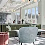 Lyst og moderne hotellobbyområde med en grønn fløyelsofa, lenestoler og store vinduer som slipper inn sollys.