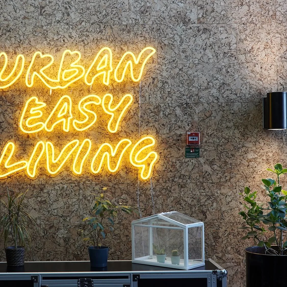 Neon sign reads "URBAN EASY LIVING" on a cork wall, flanked by plants and wall lights.