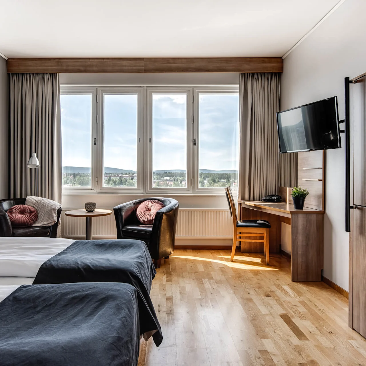 Ett prydligt hotellrum med två sängar med grå sängkläder, två fåtöljer vid ett fönster, en väggmonterad TV och ett skrivbord med en stol med utsikt över en naturskön vy.