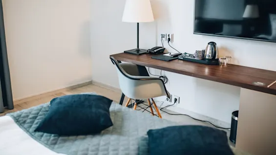 Et skrivebord med lampe, telefon og elkedel står mod en væg under et monteret TV i et hotelværelse. En seng med blå puder er synlig i forgrunden.