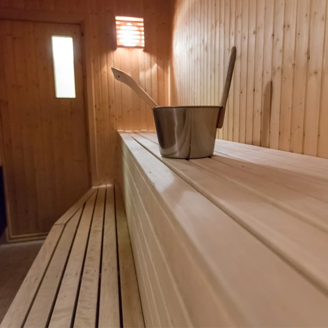 A wooden sauna has a metal bucket and ladle on a bench, with a warm light and a closed wooden door in the background. 