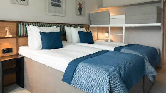 Et koselig hotellrom med to enkeltsenger og en innebygd køyeseng, med blå og hvite sengetøy og varm belysning.