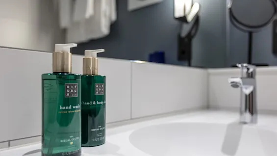 Two green bottles labeled "hand wash" and "hand & body lotion" on a bathroom counter with a sink and mirror in the background.