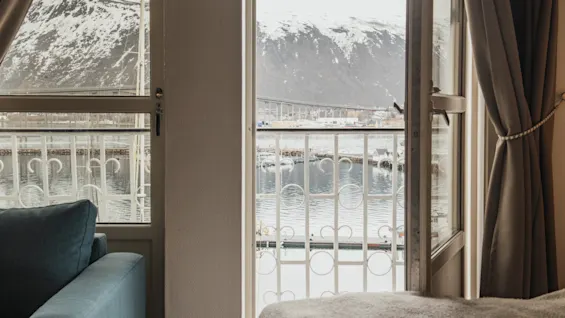 A cozy room with a balcony view of a snowy mountain landscape, a body of water, and a distant bridge.