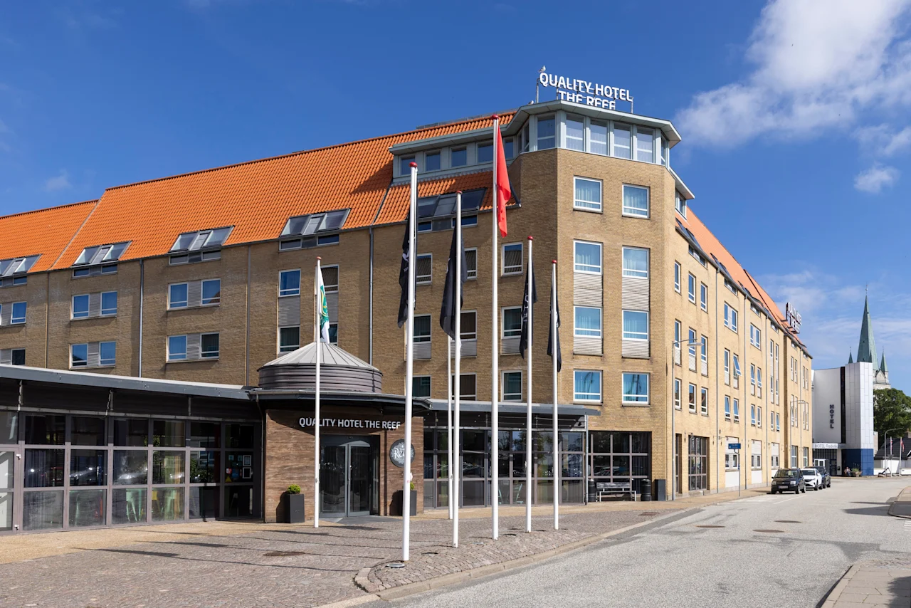 Et fleretagers hotel med rødt tegltag står på en solrig gade, med skilte der viser "QUALITY HOTEL THE REEF" på facaden. Flere flag er placeret foran.