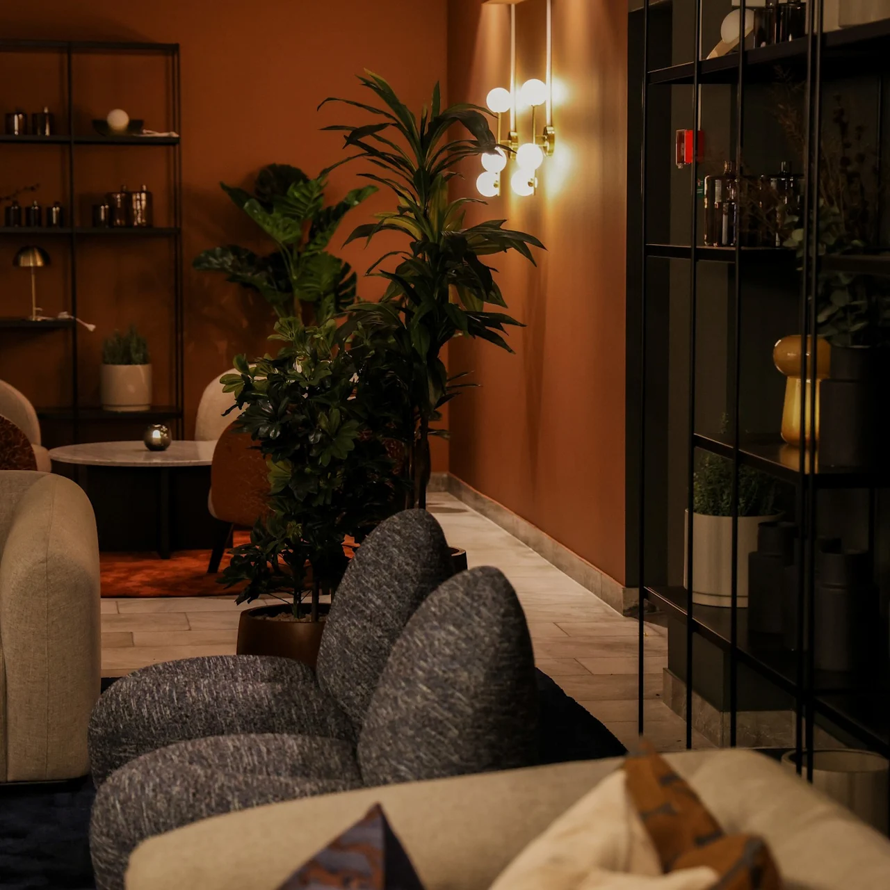 Couches and armchairs are arranged around small tables in a warmly lit, modern living room with plants and decorative shelving.