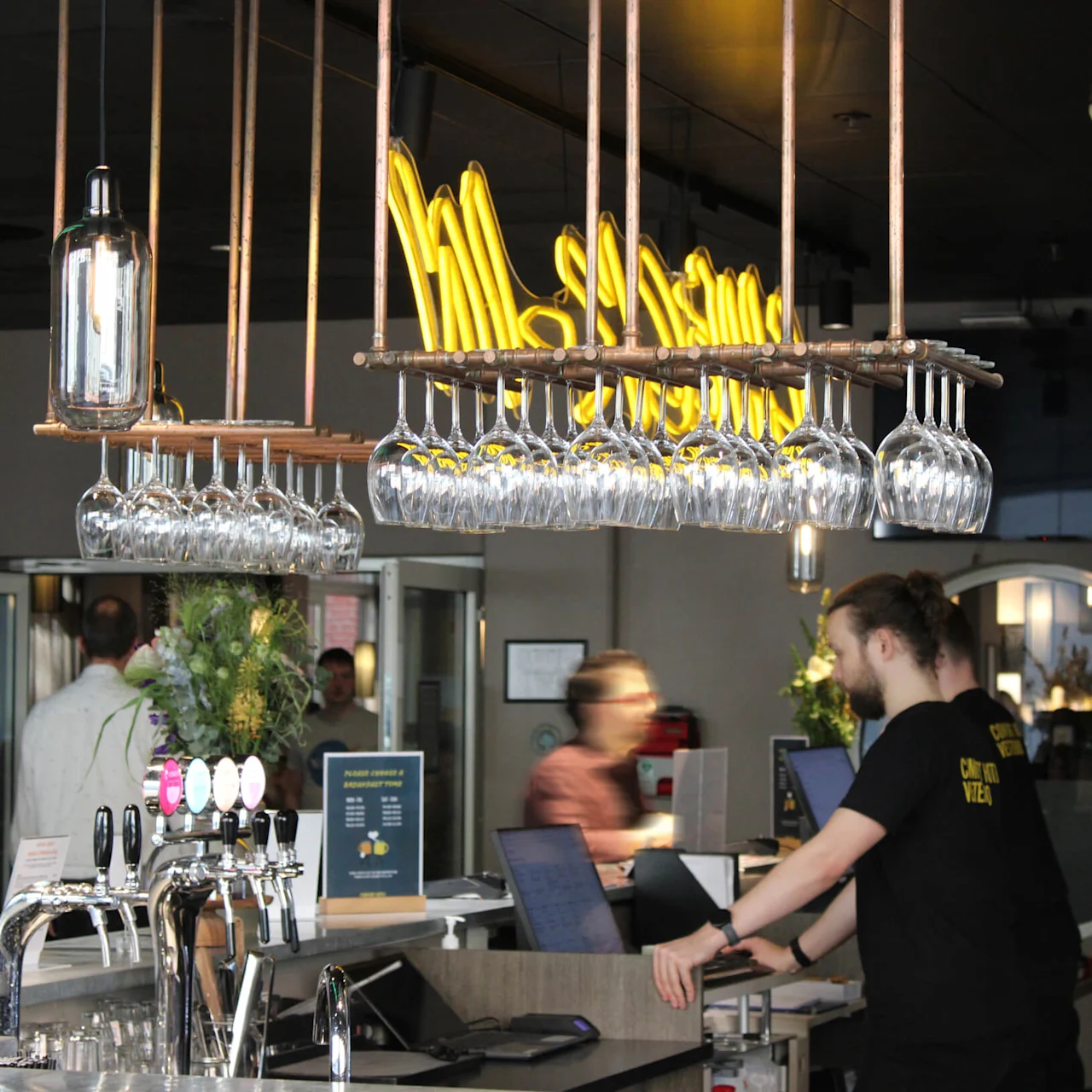 En bar med opphengte vinglass, tappetårn for øl og en bartender ved en datamaskin. Et gult neonskilt er delvis synlig i bakgrunnen.