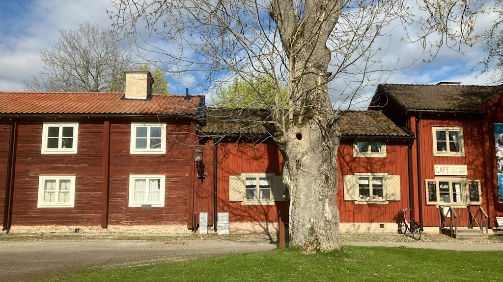 Red wooden buildings with a cafe sign and a large tree in Wadköping in Örebro. Text reads CAFE and TAKE AWAY.