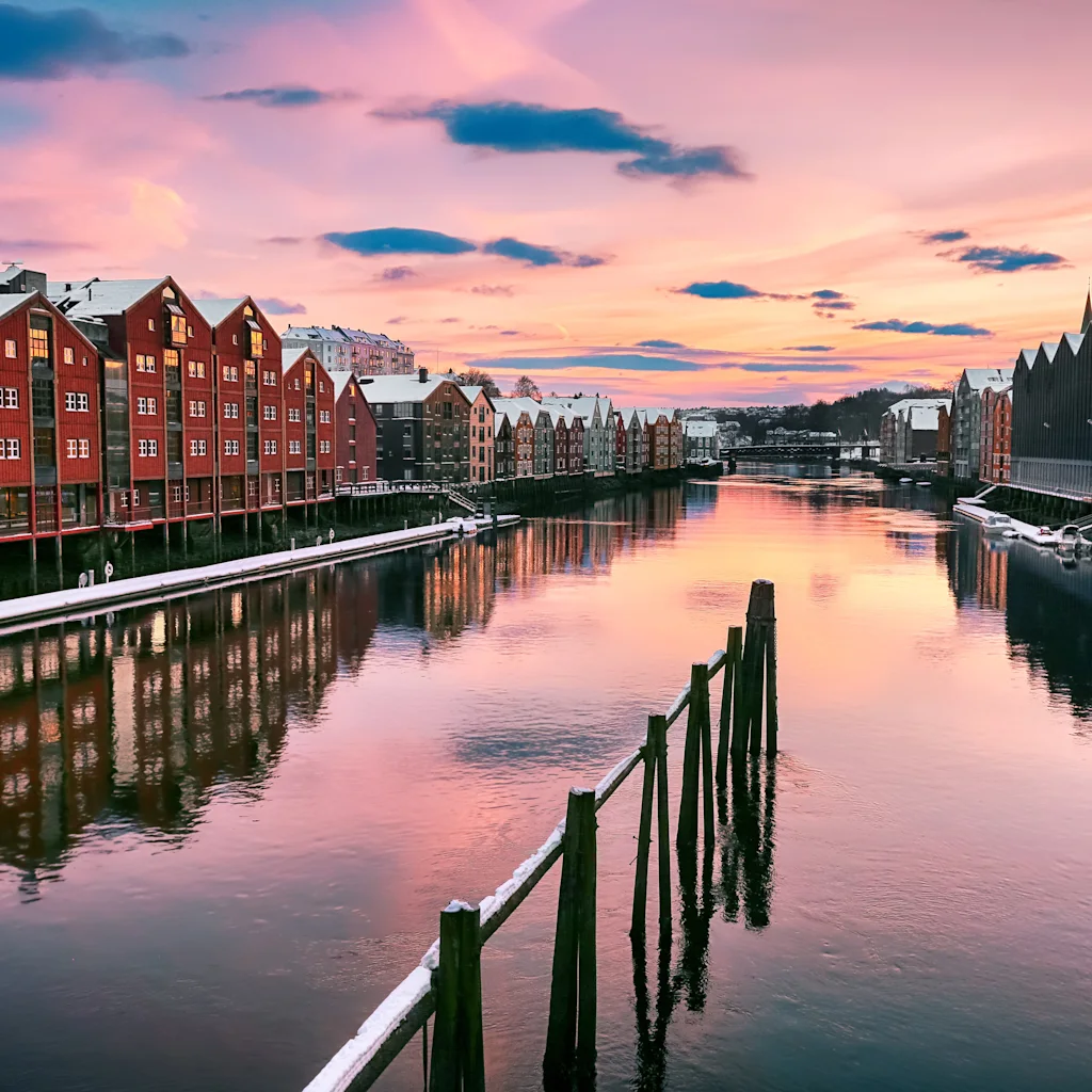 Trondheims stadsbild vid solnedgång/soluppgång. Färgglada byggnader längs en snötäckt flod speglar den rosa och lila himlen.