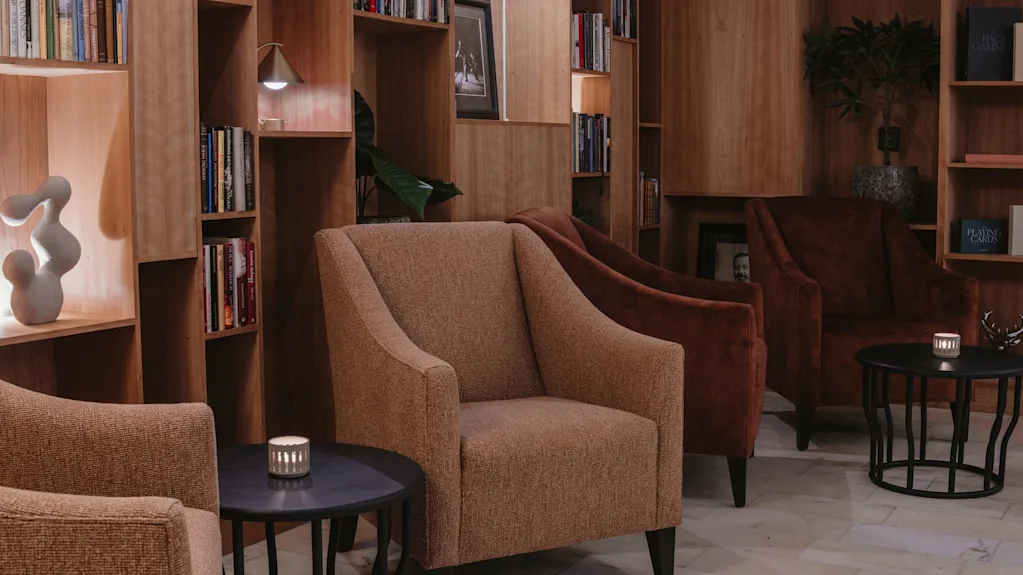 En varm och inbjudande hotellounge med bekväma sittplatser, bokhyllor och mjuk belysning. En bok med titeln Playing Cards är synlig.