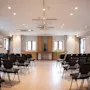 Rows of empty black chairs face a wooden podium with a vase, in a bright room with large windows and framed pictures on light-colored walls.