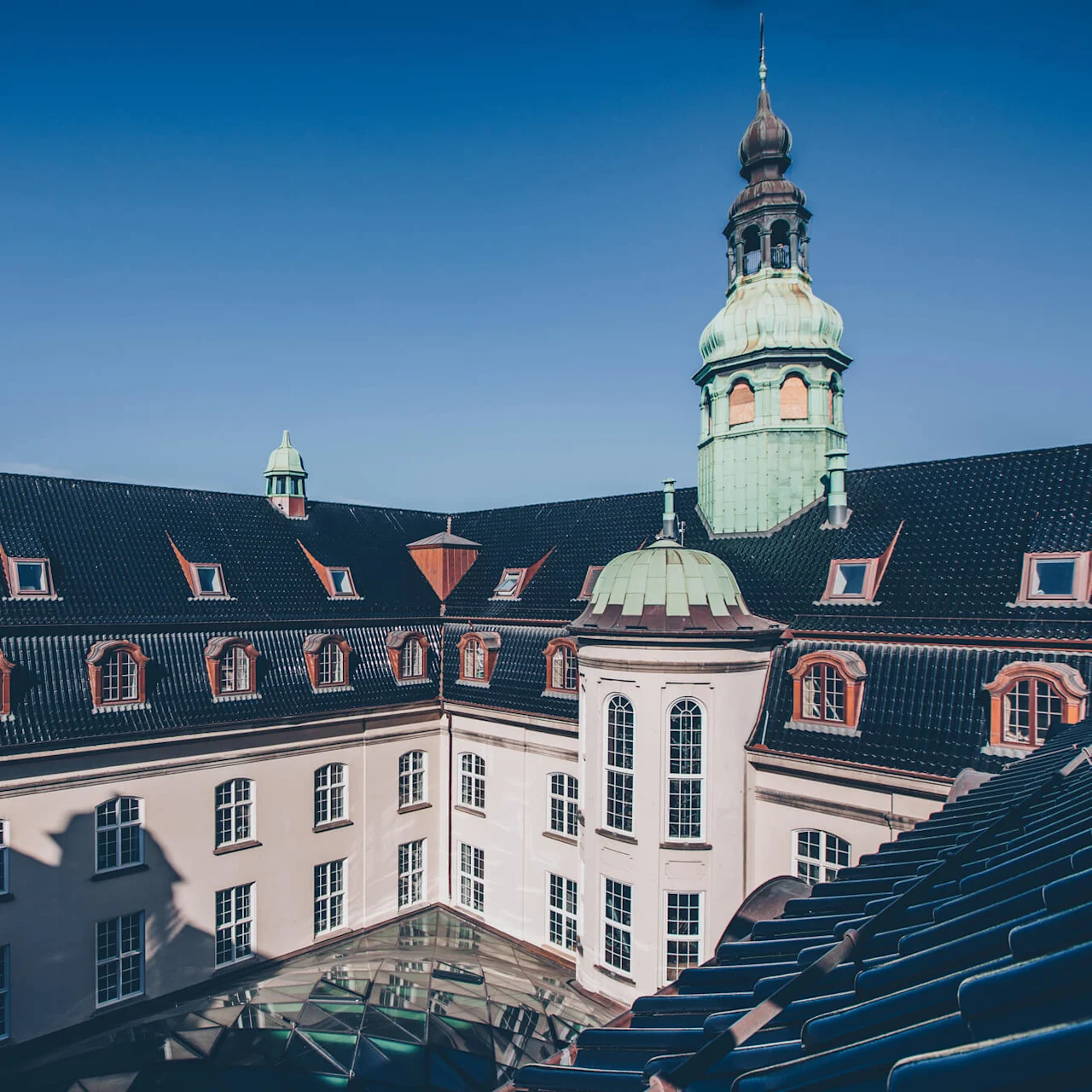 An ornate historical building displays a green dome and tower against a clear blue sky, surrounded by multiple windows and a dark, tiled roof.