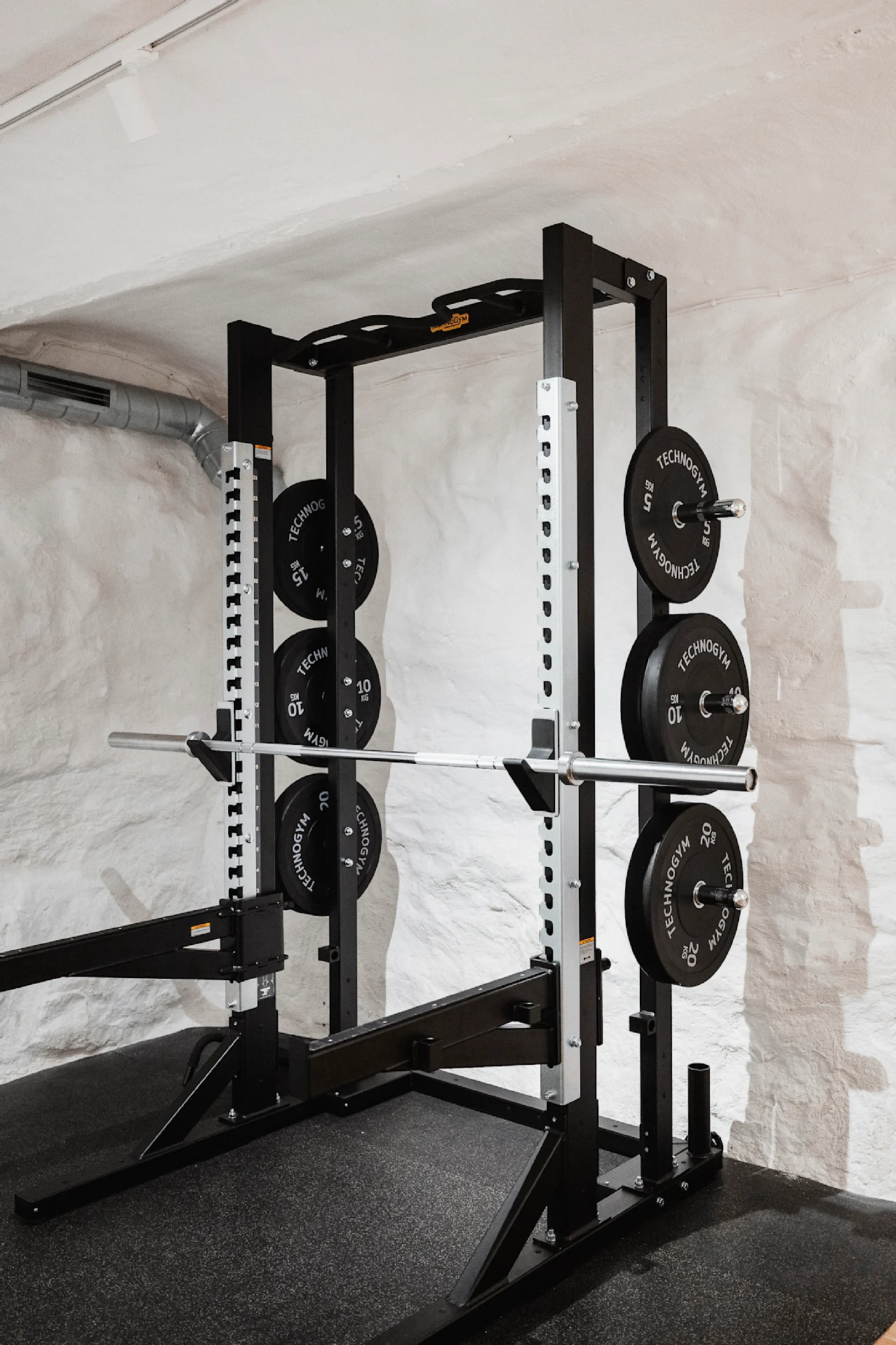 Et vægtløftestativ holder en vægtstang med forskellige vægtskiver mærket "TECHNOGYM" mod en hvid murstensvæg i et træningscenter.